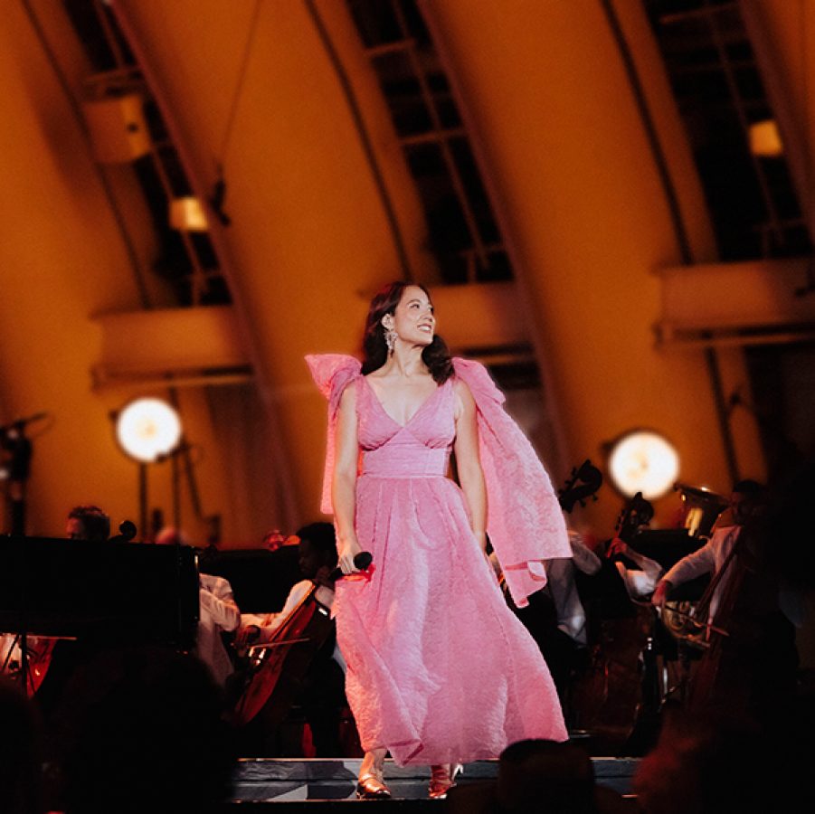 Singer Laufey stands on a softly lit stage while wearing a pink evening gown and holding a microphone. Behind her is an orchestra. 